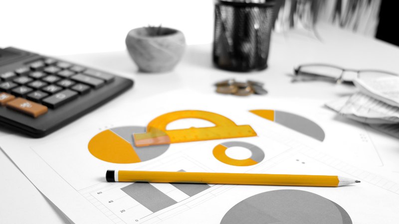 A desk calculator, printed charts, a yellow pencil and reading glasses laid out on a work table